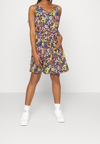 Květinové tištěné bezramínkové šaty s véčkovým výstřihem, přestřiženým pasem a sukní ve tvaru A, zdobené jasnými barvami na tmavém pozadí. Na sobě bílé tenisky.