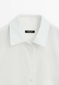 Chemise blanche à col avec boutons et une étiquette noire visible indiquant « Massimo Dutti Made in China ».