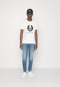 Baltas īsām piedurknēm t-krekls ar melnu "Belstaff" logo un ērgļa dizainu, apvienots ar gaiši ziliem šauriem džinsiem un baltiem sporta apaviem, modele stāv.