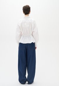 Camicia bianca leggera con dettagli arricciati in vita, abbinata a pantaloni in denim blu scuro a taglio morbido, visti da dietro.
