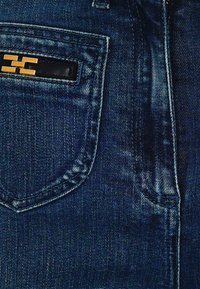 Jeans in denim blu scuro con una tasca frontale caratterizzata da un dettaglio logo nero e oro. Il tessuto presenta una leggera trama e una leggera scoloritura.