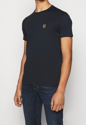 Mand iført en simpel sort t-shirt med korte ærmer og et lille rektangulært logo på brystet samt mørkeblå jeans, stående foran en hvid baggrund.