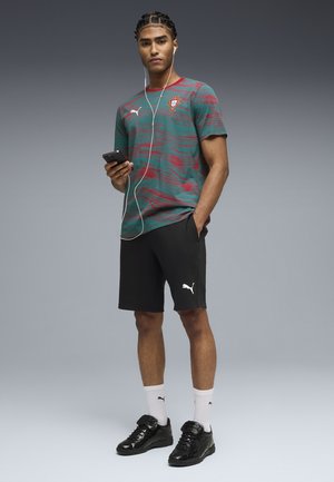 Młody mężczyzna z warkoczami, ubrany w koszulkę piłkarską Portugalii i czarne spodenki Puma, trzymający telefon z podłączonymi słuchawkami.