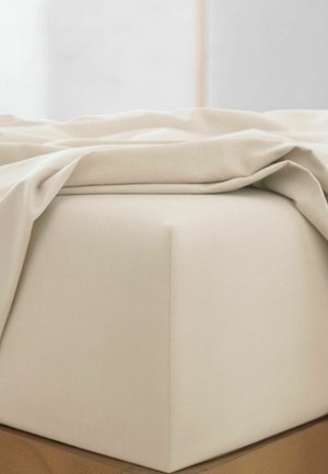 Nærbillede af et beige formsyet lagen på en madras med et delvist foldet beige top lagen på en træramme til seng.