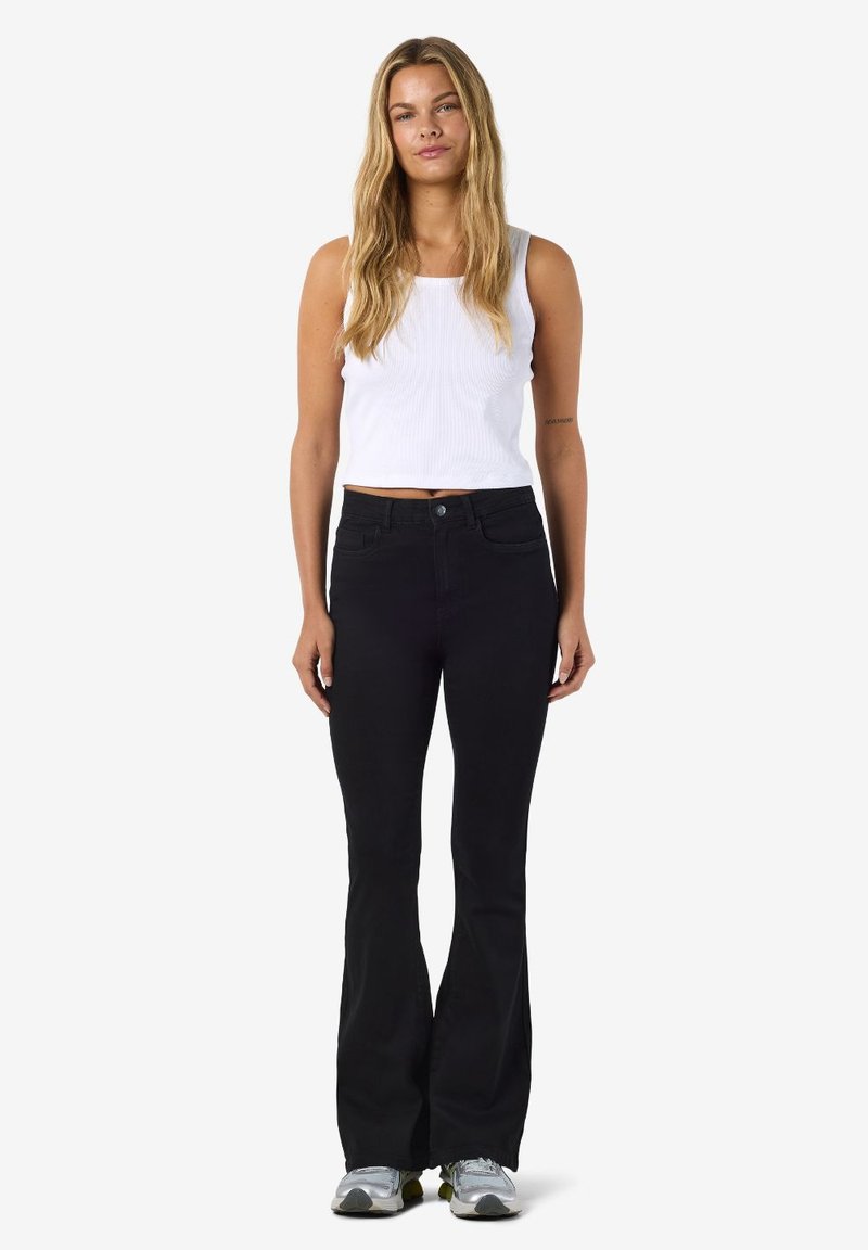 Crne trapez traperice s visokim strukom, izrađene od trapera. Uparene s bijelom rebrastom majicom bez rukava. Glatka tekstura, minimalistički dizajn.