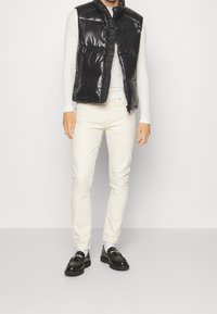 Schwarze Pufferweste mit glänzender Oberfläche, Reißverschluss und hohem Kragen, getragen über einem weißen Langarmshirt und kombiniert mit hellen Hosen und schwarzen Loafers.