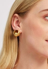 Zelta apļveida auskari ar gludu, spīdīgu virsmu un skulpturālu dizainu. Nēsāti modeļa ausī, daļēji redzami mati.