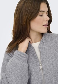 Veste aviateur grise zippée en tissu texturé, avec poignets et col côtelés, doublure intérieure lisse visible au niveau de l'encolure.