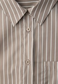 Chemise beige avec des rayures blanches verticales, un col, deux boutons et une poche sur le côté droit.