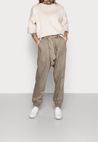 Světle béžová oversized mikina a volné taupe kalhoty s uvázaným pasem, doplněné bílými teniskami, na neutrálním pozadí.