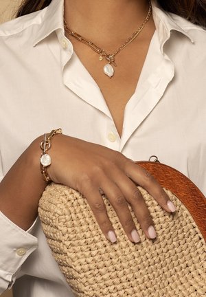 Gouden ketting met een witte parel als accent, bijpassende gouden armband en een geweven stro clutch met een houten sluiting. Witte overhemd achtergrond.