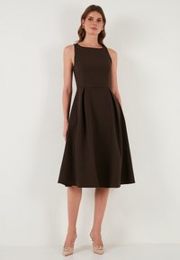Ciemnobrązowa sukienka bez rękawów z dopasowanym górnym fasonem i rozkloszowanym dołem. Gładki materiał z fałdami, w parze z beżowymi szpilkami.