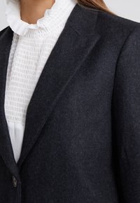 Blazer di lana grigio scuro con rever a notched, due bottoni e una texture liscia, indossato sopra una blusa bianca con rouches e texture. 