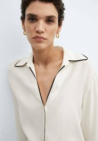 Cremfarbene Bluse mit schwarzem Piping, kragenförmigem Halsauschnitt und einem lässigen Schnitt. Glattes Material mit subtiler Schimmerwirkung, präsentiert an einem Model mit Creolen.