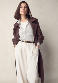 Brun mockatrenchcoat över randig skjorta med knappar, kremfärgade byxor med hög midja, med bälte och gulddetaljer. Modellen poserar med händerna i fickorna.