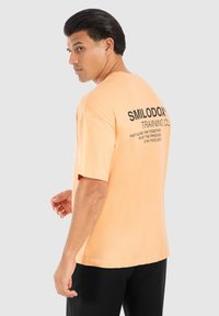 Kortærmet t-shirt i pastelfarvet orange bomuld, med sort tekst på ryggen. Lige snit, afslappet pasform, minimalistisk design.