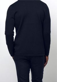 Person står med händerna i fickorna iförd en långärmad svart tröja och svarta byxor, sedd bakifrån mot en neutral bakgrund.