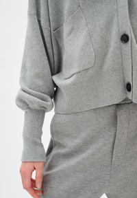 Gray cardigan made of soft material, featuring a cropped fit, rolled sleeves, two front pockets, and black buttons. Paired with matching gray pants.