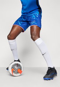 Nike kék focisort hullámos mintával, narancssárga részletekkel, fekete stoplis cipővel és fehér térdharisnyával párosítva, egy fekete-fehér focilabda mellett.