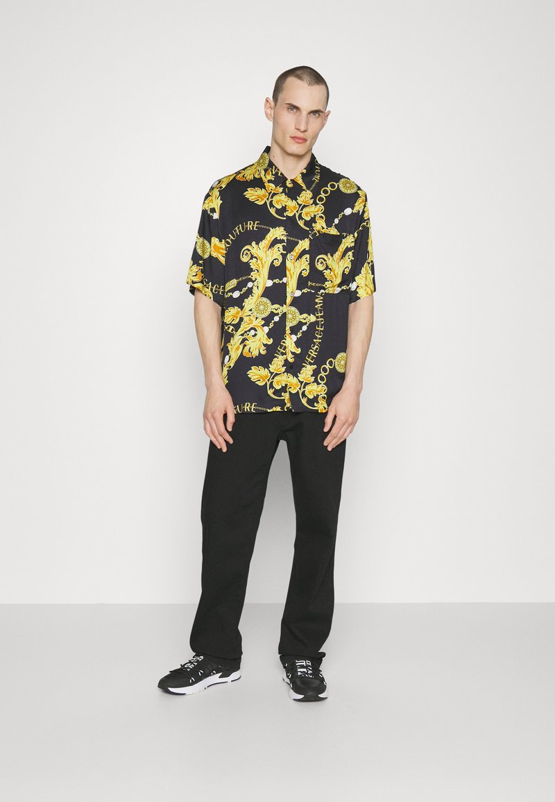 Chemise noire à manches courtes avec imprimé baroque jaune et texte, associée à un pantalon noir et des baskets noires. Le design présente une coupe ample.