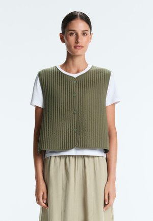 Femme aux cheveux foncés coiffés en chignon bas, portant un gilet matelassé vert olive sur un t-shirt blanc et une jupe beige, se tenant devant un fond uni.