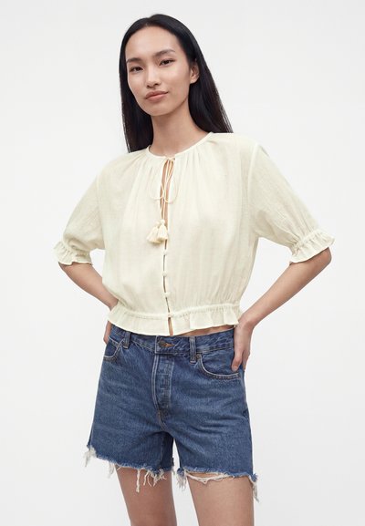 Femme aux longs cheveux foncés portant un chemisier crème avec des liens à pompons et des poignets volantés associé à un short en jean effiloché.