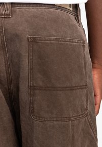 Brown cargo pants featuring a large back pocket, heavy cotton material, and reinforced stitching with a textured finish.
