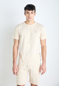 Camiseta de algodón beige con mangas cortas, escote redondo y pequeño logo bordado. Combinada con pantalones cortos beige a juego. Textura suave.