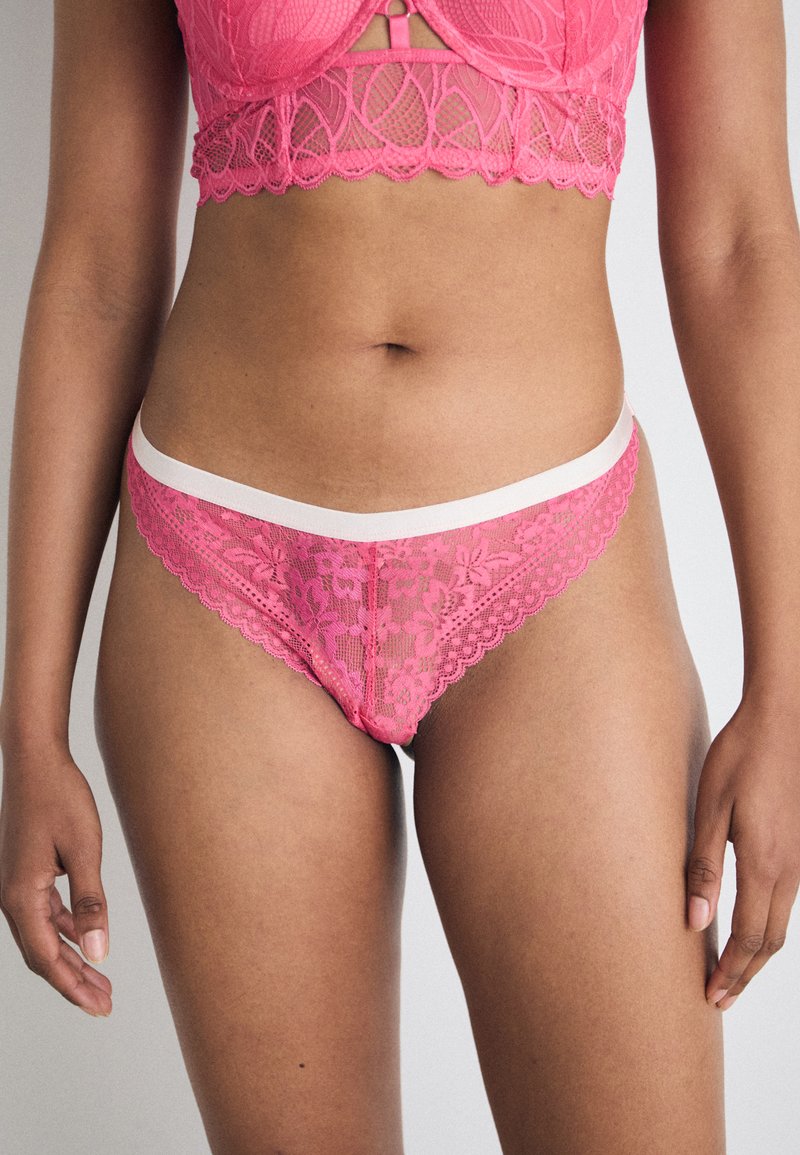 Rosa spetsstring med ett blommönster, med vågiga kanter och ett vitt midjeband. Mjuk textur och delikata design detaljer.
