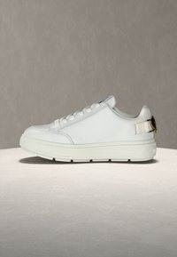 Λευκό χαμηλό αθλητικό παπούτσι με χοντρή σόλα, μπροστινό δέσιμο με κορδόνια και χρυσή αγκράφα στο πίσω μέρος, τοποθετημένο σε ουδέτερη επιφάνεια.