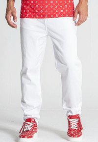 Pantaloni in denim bianco con taglio dritto e cuciture visibili. Abbinati a sneakers rosse con motivo e lacci bianchi, dotate di suole in gomma.