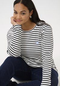 T-shirt à manches longues rayé blanc et noir, orné d'un patch en forme de cœur bleu clair. Tissu doux avec un col rond et une coupe décontractée.