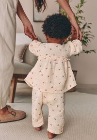 Tenue crème légèrement texturée avec des motifs de cerises rouges ; manches bouffantes sur la chemise, associée à un pantalon assorti ; l'enfant est debout.
