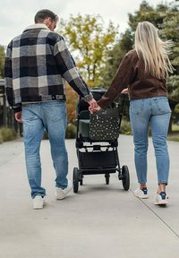 Černo-bílá károvaná bunda, hnědý svetr, modré džíny; černý kočárek se zelenou látkovou taškou s barevnými vzory, venkovní prostředí.