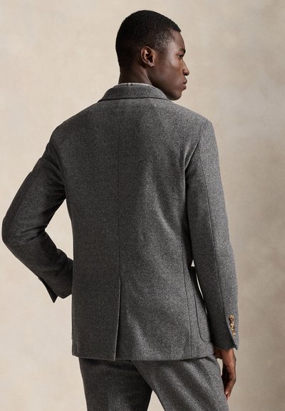 Man in een grijs wollen colbert en bijpassende broek staat met zijn rug naar de camera, kijkt opzij tegen een neutrale beige achtergrond.