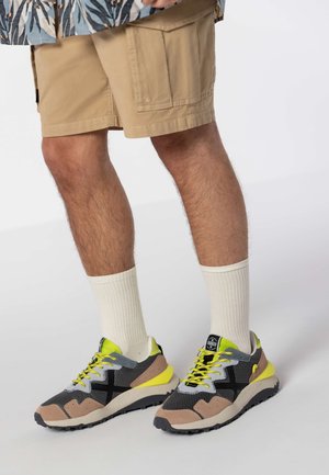 Sneakers hebben een combinatie van zwart gaas en beige suède, met gele accenten en grijze veters. Draag ze met crème sokken en kaki shorts.