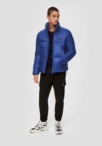 Blauwe pufferjack met een hoge kraag, gecombineerd met zwarte cargo-broeken en witte sneakers. Beschikt over een logo-opdruk en een gewatteerde textuur.