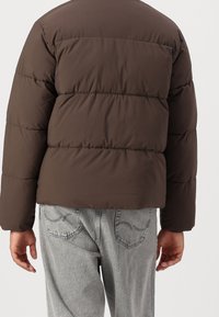 Hnědá prošívaná bunda s volným střihem, která má prošívané části a hladkou texturu. Kombinovaná s světle šedými džínovými kalhotami.