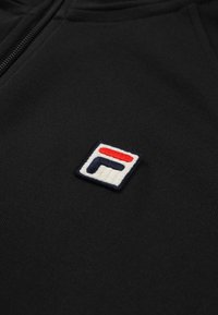 Svart tyghuva med en framträdande broderad rektangulär logotyp i blått, vitt och rött med en klassisk blockdesign och texturerad yta.