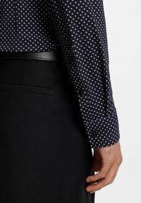 Braccio e mano di un uomo che indossa una camicia a maniche lunghe con motivo blu navy infilata in pantaloni neri con una cintura nera, su uno sfondo bianco.