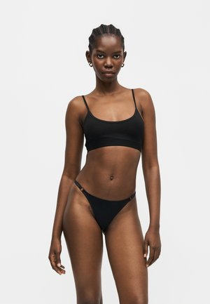 Mujer con el cabello trenzado que lleva un top corto negro con tirantes finos y tanga negro a juego, de pie frente a un fondo claro y liso.