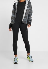 Chaqueta de camuflaje negra con un forro interior ligero, camiseta negra, pantalones negros ajustados con bolsillos con cremallera y zapatillas deportivas azul-verde.