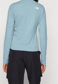 Haut à manches longues bleu clair avec un tissu texturé, arborant un petit logo blanc dans le haut du dos et un design ajusté. Associé à un pantalon cargo noir.