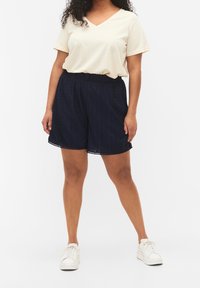 Pantaloni corte blu marino in tessuto texturizzato con righe verticali e orlo a scaglie. Abbinati a una t-shirt beige chiaro con scollatura a V e scarpe da ginnastica bianche.