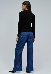 Suéter negro con detalles de botones dorados, combinado con jeans azul de pierna ancha de cintura alta. Tacones negros completan el atuendo.