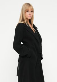 Mulher loira com cabelo liso, vestindo um longo casaco preto e com as mãos nos bolsos, em pé contra um fundo branco.