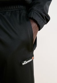 Main d'une personne dans la poche d'un pantalon noir ellesse avec un écusson logo visible sur la cuisse, sur un fond uni.