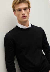 Schwarzer Strickpullover mit Rundhalsausschnitt, getragen über einem weißen Hemd. Der Stoff wirkt glatt, mit einer figurbetonten Passform und langen Ärmeln.