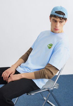 Νέος άνδρας με ανοιχτό μπλε καπέλο και πουκάμισο φορεμένα πάνω από καφέ μακριά μανίκια, κάθεται σε μεταλλική αναδιπλούμενη καρέκλα, κοιτάζοντας προς τα δεξιά του πάνω σε μπλε χαλί.