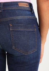 Vaqueros de mezclilla azul oscuro con cintura alta, costuras naranjas y un bolsillo trasero texturizado con una etiqueta de marca en la cintura.